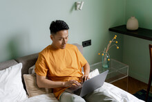 A man uses a laptop while sitting on a bed