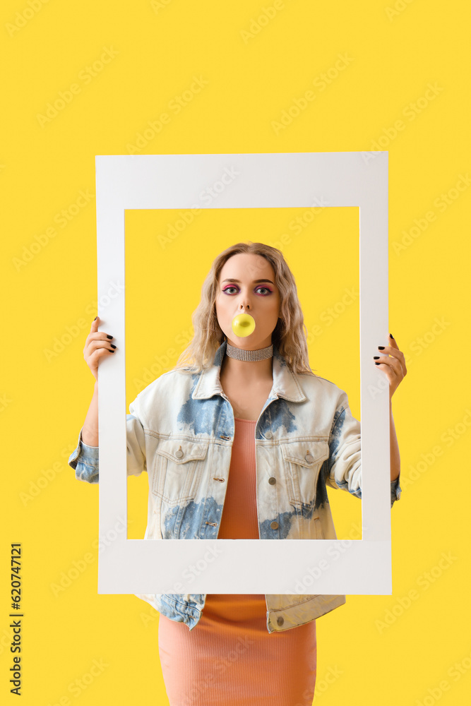 Young woman with frame chewing gum on yellow background