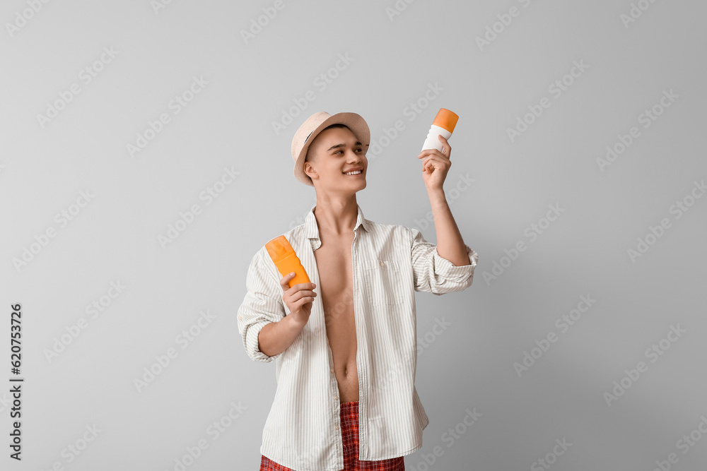 Happy young man with different bottles of sunscreen cream on grey background