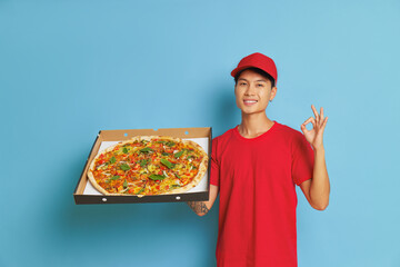 Wall Mural - Cheerful boy with box with large pizza in one hand on blue background shows ok sign with other hand, wearing red cap and red t-shirt, fast delivery concept, copy space