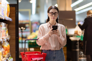 Wall Mural - Portrait of young serious Caucasian woman wearing glasses holds shopping basket and uses smartphone. Online shopping and consumerism