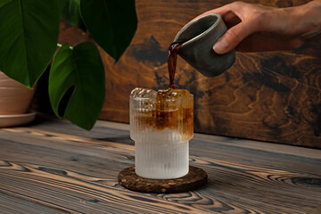 Wall Mural - Espresso tonic cold refreshing drink in the making. Pouring coffee into a glass filled with ice cubes and soda water, on wooden background