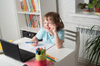 © Anastasia - Happy girl sits at a table with a laptop and books, looks into the laptop and smiles. Children learning at home. Distance learning online education. Back to school.