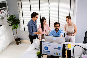 Cheerful young group of Asian businessmen in casual with one sitting and pointing at the computer display and colleagues discussing around him during the meeting. Diversity of businessmen in a meeting