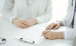 © rogerphoto - Doctor and patient sitting near of each other at the desk in clinic. The focus is on female physician's hands filling up the medication history record form or checklist, close up. Medicine concept