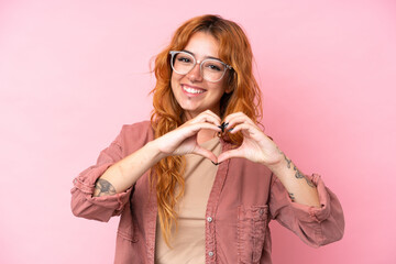 Wall Mural - young caucasian woman isolated on pink background with glasses making heart with hands