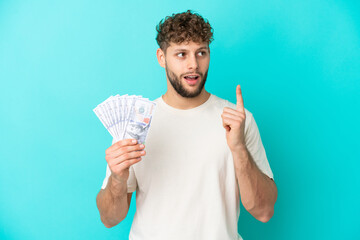 Canvas Print - Young caucasian man taking a lot of money isolated on blue background thinking an idea pointing the finger up