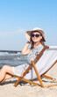 © rogerphoto - Happy brunette woman wearing sunglasses and hat relaxing on a wooden deck chair at the ocean beach