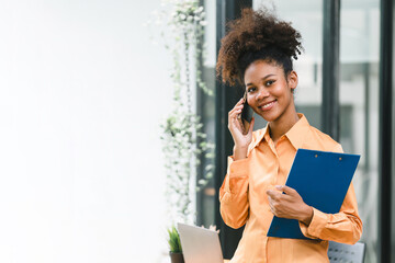 Portrait of a young, smiling, and cheerful African American businesswoman working in a casual office, holding a financial report folder while making a business call.