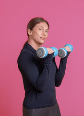 Wall Mural - Workout concept. Young athletic woman in sportswear trains biceps muscles with dumbbells in her hands on pink background. Bodybuilding and fitness. Healthy lifestyle