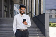 © Liubomir - Closeup businessman walking city and using application on smartphone, a smiling Hispanic man holding a phone in his hands, browsing online pages, typing messages, and choosing in online store.