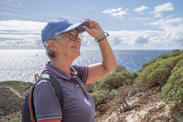 Sticker - Active senior woman in blue hat walking on trail outdoors on country excursion. Scenic seascape on background. Happy caucasian woman enjoying travel or retirement leading healthy lifestyle.