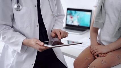 Wall Mural - Doctor and kid patient sitting opposite each other in clinic, close up. Unknown female physician using tablet computer near little girl. Medicine concept