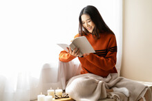 Beautiful Young Asian Woman Reading Book Wearing Knitted Warm Sweater Sit On Windowsill In Room Decorated With Candle At Home. Korea Or Japanese Girl Relaxing At Home. Cozy Lifestyle, Hygge Concept
