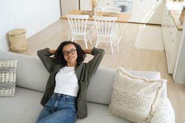 Sticker - Relaxed girl in glasses dreams on cozy couch at home. Calm woman enjoys lazy break, resting with hands behind head.