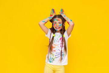 Wall Mural - Portrait of a girl with a face painted with multicolored paints. Children's creativity. The child shows his ears above his head. Yellow isolated background.