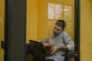 Close up shot of good looking male person working on his lap top in phone booth at the office while having a phone call