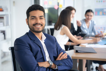 Attractive asian young confident businessman sitting at the office table with group of colleagues in the background. Business man with coworkers in meeting room.