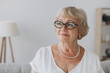 Smiling senior blonde haired woman looking at camera, happy old lady in glasses posing at home indoor, positive single senior retired female sitting on sofa in living room headshot portrait