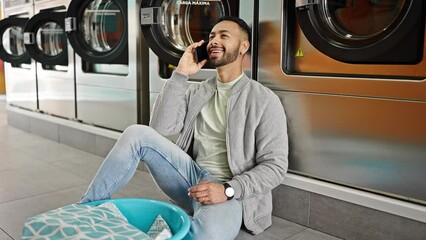 Wall Mural - Young hispanic man speaking on the phone sitting on the floor at laundry facility