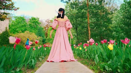 Wall Mural - Fantasy girl princess walks in blooming spring garden flowers tulips tree green grass alley. Woman queen in long royal pink dress with train puffed sleeves vintage old style. lady back rear view. 4k