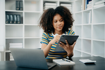 young African student studying at the college library, sitting at the desk, using a laptop computer, tablet and headphones having a video chat...