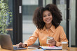 © Songsak C - Portrait of a beautiful confident businesswoman using a laptop computer holding a mobile phone sitting in a modern office. Smiling African American freelancer working online from home.
