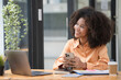 © Songsak C - Portrait of a beautiful confident businesswoman using a laptop computer holding a mobile phone sitting in a modern office. Smiling African American freelancer working online from home.