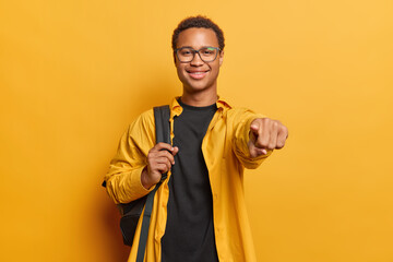 Wall Mural - Horizontal shot of handsome pleased African man points finger directly at camera chooses you to join his team carries rucksack on shoulder dressed casually isolated over vivid yellow background