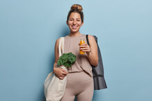 People Sport And Healthy Lifestyle Concept. Positive Young Sporty Woman Holds Bottle Of Orange Juice Carries Fabric Bag With Green Vegetables Dressed In Activewear Isolated Over Blue Background