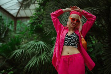 Fashionable happy smiling woman wearing trendy orange sunglasses, pink fuchsia color bandana, linen suit with oversized shirt, low waist trousers, zebra print crop top, posing in tropical garden. 