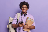 Fototapeta  - Horizontal shot of pleased curly haired Hindu student smiles gladfully uses modern texhnologies for studying and entertainment carries notepads carries backpack wears t shirt and spectacles.