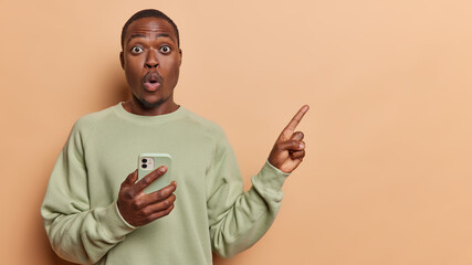 Poster - Waist up shot of African adult man pointing finger at empty space isolated on brown background recommends sale online holds mobile phone dressed in casual jumper suggests to write your text.