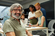 © CarlosBarquero - Portrait of an office man smiling and looking at camera. In the background there is a group of employees kipping at work. It is an indoor scene.