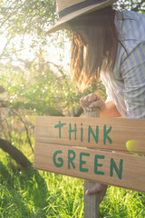 young woman with a hat in an environment of trees and sun with a wooden sign that says think green to protect the natural environment and take care of the environment
