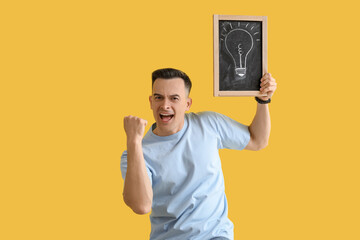 Canvas Print - Happy male teacher holding chalkboard with drawn light bulb on yellow background