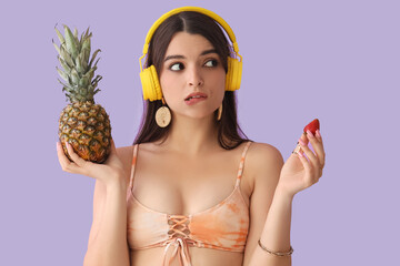 Wall Mural - Thoughtful young woman in headphones with pineapple and strawberry on lilac background