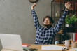 © ViDi Studio - Successful overjoyed employee business Indian man he wears casual blue checkered shirt doing winner gesture celebrate raise up hands finish job sit work at office desk with laptop pc computer indoors.