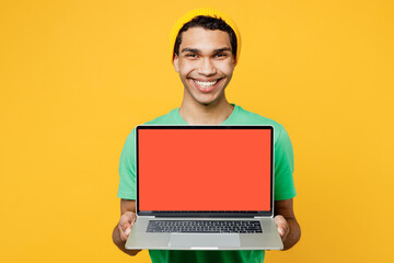 Wall Mural - Close up young IT man of African American ethnicity wears casual clothes green t-shirt hat work hold use laptop pc computer with blank screen workspace area isolated on plain yellow background studio.