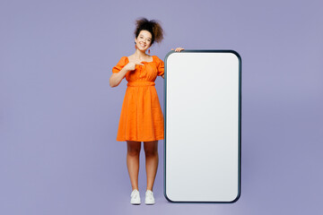 Full body young smiling latin woman she wear orange blouse casual clothes point index finger on big huge blank screen mobile cell phone smartphone with area isolated on plain purple background studio.