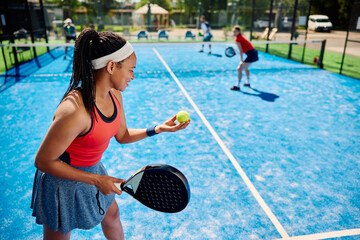 Black paddle tennis player serving ball during mixed double match on outdoor court.
