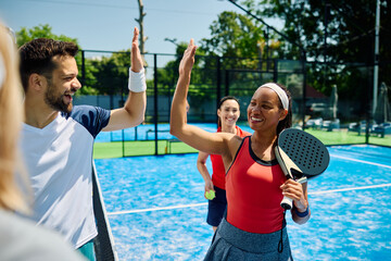 Happy black paddle tennis player giving high five to her rival after playing mixed doubles match.