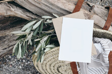 Summer Lifestyle Composition. Blank Greeting Card Invitation Mockup. French Basket, Straw Bag With Olive Tree Branches, Towel: BLurred Pebble Beach Background With Drift Wood. Mediterranean Design.