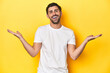 © Asier - Caucasian man in white t-shirt on yellow studio background makes scale with arms, feels happy and confident.