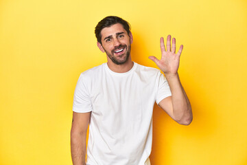 Caucasian man in white t-shirt on yellow studio background smiling cheerful showing number five with fingers.