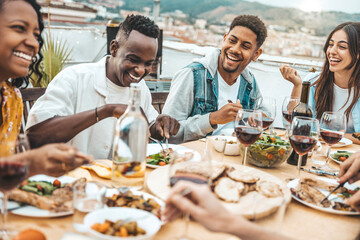 Wall Mural - Multiethnic friends having fun at rooftop bbq dinner party - Group of young people diner together sitting at restaurant dining table - Cheerful multiracial teens eating food and drinking wine outside