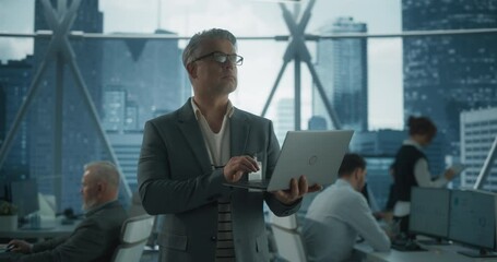 Poster - Succeful Caucasian Businessman Walking In Office With Diverse Employees In Downtown Skyscraper. Middle Aged Entreprenuer Using Laptop Computer In E-commerce Company Workspace. Dramatic Evening Edit.