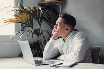 Wall Mural - Thoughtful businessman touching chin, pondering ideas or strategy, sitting at wooden work desk with laptop, freelancer working on online project, student preparing for exam at home.