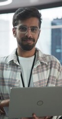 Poster - Vertical Screen: Handsome Indian Man Wearing Glasses, Smiling and Looking at Camera. Young Intelligent Male Engineer Holding a Laptop Computer. Finance Manager Working in Modern Corporate Office