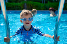 An Independent Baby Deftly Climbs A Metal Ladder In The Summer Pool After Swimming. Satisfied Child Carefully Climbs Out Of The Water Of The Summer Pool Up The Stairs Wearing Swimming Goggles
